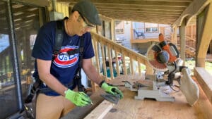 Jonathan Dunsmore measures and cuts a piece of wood in his carpentry shop. He has a safety mask and vest, along with cut-proof gloved on. There is a large saw next to him.