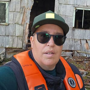 A portrait of Goox standing in front of an old cabin