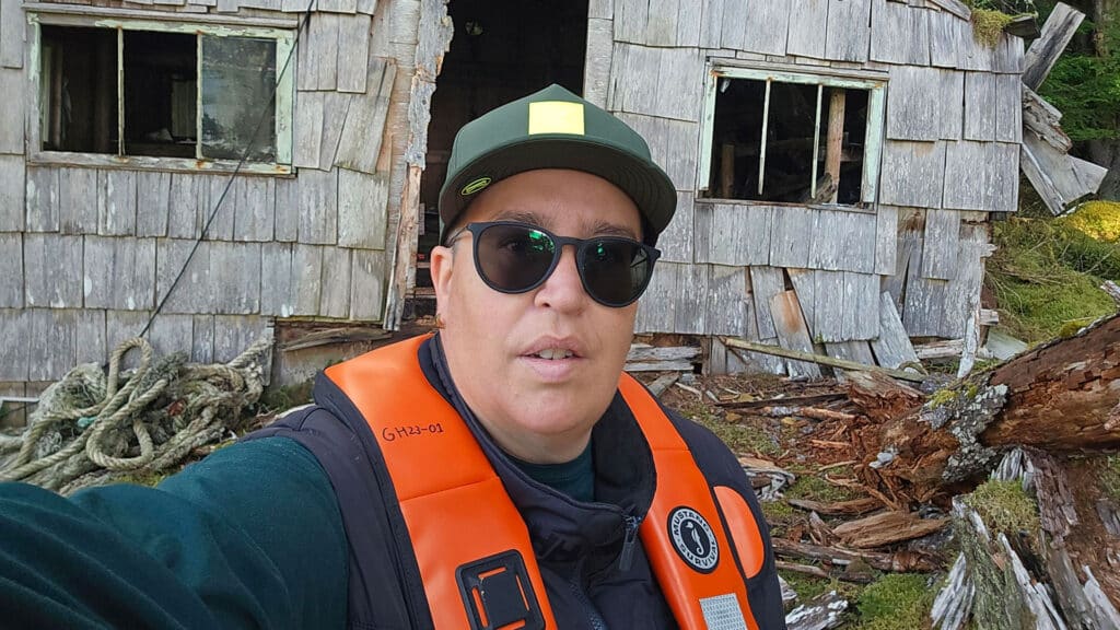 Goox stands in front of the remains of the cabin that was built to shelter those who stood the line back in 1985. The forest and moss encroach on the wooden cabin. Goox wears a hat, sunglasses, a pfd and a dark sweater.
