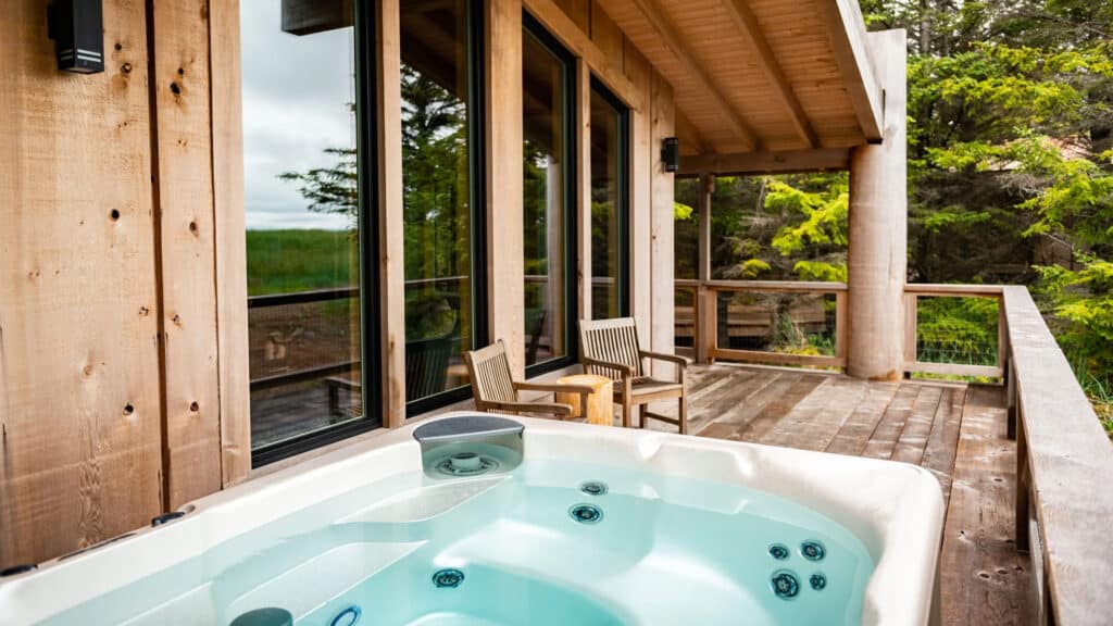 A cedar wood cabin with a large sundeck and big windows, featuring a crystal clear hot tub with the cover removed. The forest is behind, with green cedar trees.