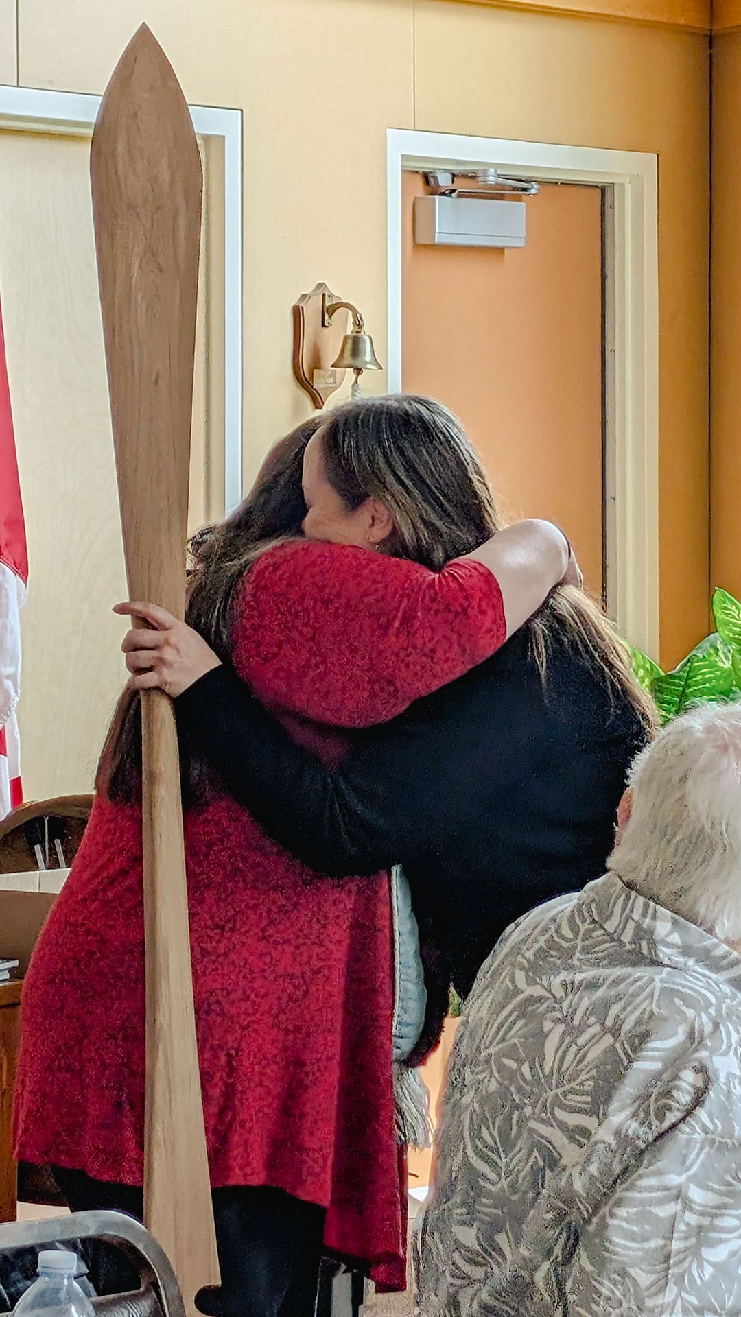 Two people hug in a room; one wears red, the other black. A tall wooden canoe paddle rests beside them.